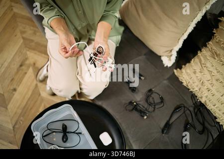 Weiße Frau mittleren Alters, die auf dem Sofa sitzt und verwickelte elektronische Kabel mit den Händen organisiert, mehrere Kabel in einen Kunststoffbehälter sortiert, Ansicht von oben zeigt Teilkörper und Arbeitsbereich Stockfoto