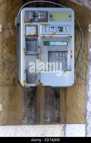 Altes, rostgesprenktes öffentliches Münztelefon, das draußen an einer verwitterten Steinmauer befestigt ist. Das abgenutzte Tastenfeld und die losen Drähte rufen Nostalgie und Verfall eines einmal-Ubiq hervor Stockfoto