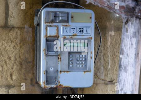 Altes, rostgesprenktes öffentliches Münztelefon, das draußen an einer verwitterten Steinmauer befestigt ist. Das abgenutzte Tastenfeld und die losen Drähte rufen Nostalgie und Verfall eines einmal-Ubiq hervor Stockfoto