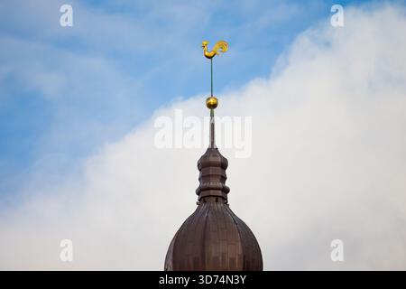 Braun gewölbte Turmspitze in Riga mit dekorativer goldener Hahn-Wetterfahne, die auf einem dünnen Turm vor hellem Himmel montiert ist und Wolkenmuster und Architektur zeigt Stockfoto