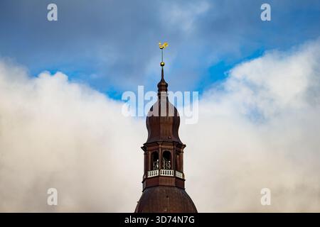 Dekorativer goldener Hahn auf einem dünnen Turm über dem braunen Kuppeldach in der Altstadt von Riga mit weichen Wolken und blauem Himmel, der einen einfachen Hintergrund um die Straße bildet Stockfoto