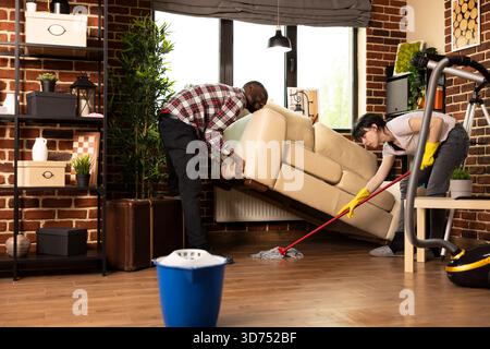 Junge, vielfältige Paare, die Hausarbeit und Hausarbeit im Wohnzimmer erledigen. Schwarzer Mann hebt die Couch an, während die Freundin den Boden mit einem Wischtuch abwischt, um Teamarbeit und Zusammenarbeit bei der Aufrechterhaltung der Sauberkeit zu Hause zu zeigen. Stockfoto
