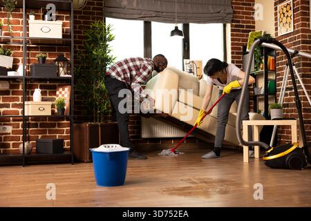 Multiethnische Ehepartner, die täglich das Wohnzimmer in einer gemütlichen, modernen Wohnung reinigen. Weiße Frau wischt den Boden ab, während ein schwarzer Mann das Sofa hebt und dabei Teamarbeit, Lebensstil und fröhliche Hausarbeit darstellt. Stockfoto