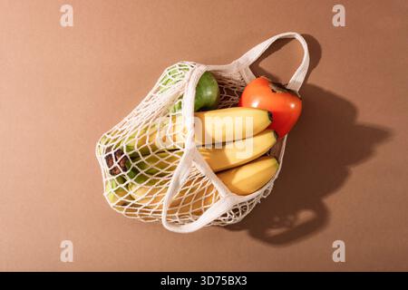 Verschiedene Früchte in einem Netzbeutel auf einer warmen Oberfläche, die eine einfache natürliche Lebensmittelanordnung mit Licht und Schatten zeigt. Draufsicht. Stockfoto