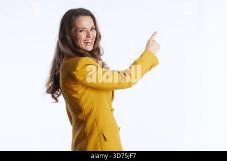 Eine lächelnde Frau mit braunem Haar, in gelbem Blazer, zeigt den rechten Zeigefinger nach oben und rechts. Vor weißem Hintergrund überträgt sie Richtung Stockfoto