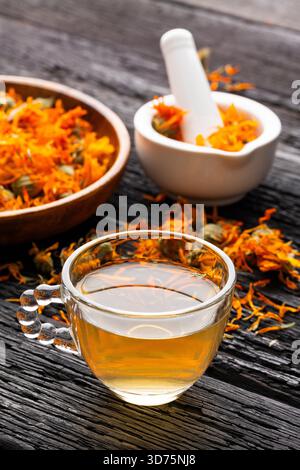 Eine Tasse Calendula-Tee sitzt auf einem rustikalen Holztisch, begleitet von getrockneten Blütenblättern und einem weißen Mörtel mit Blumen. Stockfoto
