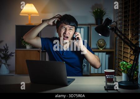 Gestresster Mann, der nachts mit dem Smartphone spricht, während er mit einem Laptop arbeitet Stockfoto