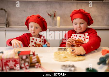 Zwei fröhliche Kinder in roten Kochmützen und Schürzen verwenden Keksschneider, um Teig auf einem Tisch in einer warmen Küche zu Formen. Die Szene fängt den Spaß eines Urlaubs ein Stockfoto