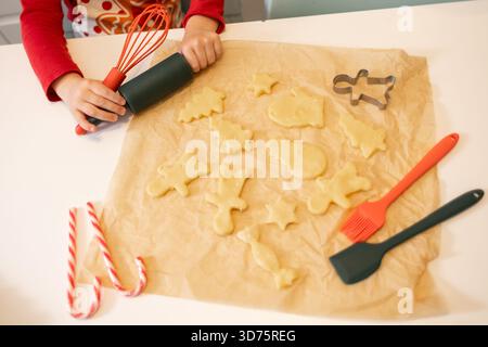 Ein Kind rollt Teig für Weihnachtskekse aus, umgeben von verschiedenen Ausstechern und Backwerkzeugen. Süßigkeiten sind ebenfalls vorhanden, was einen festlichen Tu hinzufügt Stockfoto