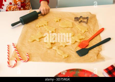 Ein Kind rollt Keksteig in einer gemütlichen Küche aus und bereitet festliche Formen wie Sterne und Lebkuchen zu. Zuckerstangen und Dekorationswerkzeuge ergänzen den Urlaub Stockfoto