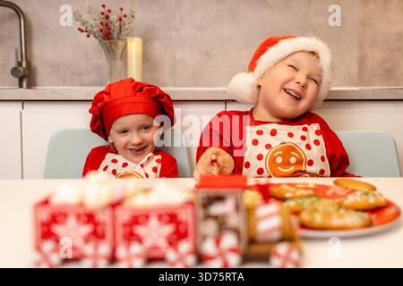 Zwei fröhliche Kinder in festlichen Outfits lachen, während sie an einem Tisch mit dekorierten Keksen in einer gemütlichen Küche sitzen. Sie feiern die Stockfoto