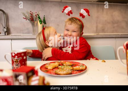 Zwei fröhliche Kinder umarmen sich, während sie in einer warmen Küche Weihnachtsschmuck tragen. Sie sind umgeben von bunten Keksen und dekorierten Tassen, die die teilen Stockfoto