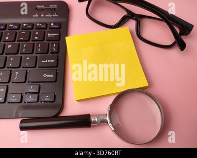 Das Bild zeigt eine Vielzahl von Büroelementen, darunter eine Tastatur, einen Stapel gelber Haftnotizen, eine Brille und eine Lupe. Stockfoto