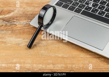 Neben einem Laptop befindet sich eine Lupe auf einem Holztisch. Die Szene schlägt eine Umgebung für Fokus, Untersuchung oder Detailarbeit vor. Stockfoto