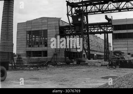 Slawjansk, Region Donezk, ukrainische SSR, Sowjetunion. 1960er Jahre Bau einer neuen Glaswerkstatt in der Artem Armature-Isolator Factory (AIZ). Ein massiver Portalkran steht im Vordergrund in der Nähe von Stapeln von Betonplatten. Das unvollendete Stahlbetongebäude verfügt über große Fensteröffnungen. Links ist ein alter gemauerter Schornstein zu sehen. Dieses archivierte Schwarzweiß-Foto zeigt den industriellen Expansions- und Bauboom in der friedlichen Donbass-Region lange vor dem Krieg. Stockfoto