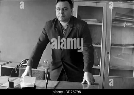 Slawjansk, Region Donezk, ukrainische SSR, Sowjetunion. Ende der 1960er Jahre Ein Porträt eines Managers oder Chefingenieurs, der an seinem Schreibtisch in einem Büro im AIZ-Werk steht. Der Mann in einer dunklen Jacke beugt sich mit den Händen auf den Tisch und blickt ernsthaft in die Kamera. Ein Schrank mit Dokumenten und ein Telefon sind im Hintergrund sichtbar. Dieses archivierte Schwarzweiß-Foto zeigt das Bild eines Industriellen Führers und einer Verwaltungsarbeit in der friedlichen Donbass-Region lange vor dem Krieg. Stockfoto