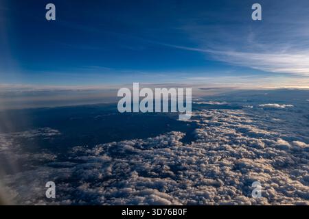 Hohe Luftaufnahme aus einem Flugzeug, das ein riesiges Meer aus weißen, flauschigen Wolken zeigt, das sich über eine ferne Landschaft erstreckt und vom warmen Sonnenlicht beleuchtet wird Stockfoto