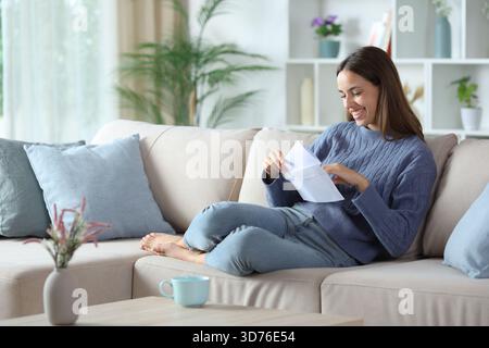 Glückliche Frau in Blau, die einen Brief in einen Umschlag legt und zu Hause auf einer Couch sitzt Stockfoto