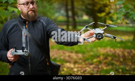 Ein bärtiger Mann in einer schwarzen Jacke steht draußen und zeigt eine kleine Drohne, während er eine Fernbedienung hält. Der umliegende Park bietet farbenfrohe Herbstblätter. Stockfoto