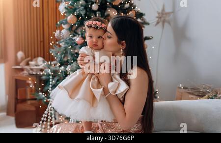 Ein freudiger Moment wird festgehalten, als eine Mutter ihr kleines Mädchen, das in einem wunderschönen Outfit gekleidet ist, vor einem glitzernden Weihnachtsbaum liebevoll küsst. Stockfoto