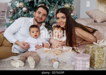 Eine Familie genießt einen festlichen Moment zusammen mit einem wunderschön geschmückten Weihnachtsbaum. Zwei kleine Kinder sitzen mit ihren Eltern, umgeben von G Stockfoto