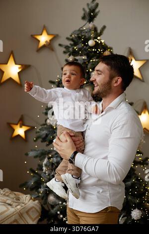 Ein fröhlicher Vater hält seinen Sohn vor einem wunderschön geschmückten Weihnachtsbaum. Sie teilen einen besonderen Moment mit Lichtern und Sternen Stockfoto