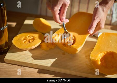 Nahaufnahme von Hand, die Samen von Kürbis mit Löffel auf Holzbrett in der Küche aufnimmt. Person in Schürze, die Kürbis zum Kochen vorbereitet Stockfoto