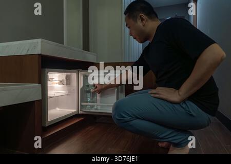 Ein Mann öffnet einen kleinen Kühlschrank, der in einem Holzschrank in einem dunklen Hotelzimmer eingebaut ist, und das warme Innenlicht steht im Kontrast zur dunklen Umgebung. Stockfoto