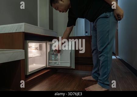 Ein Mann öffnet einen kleinen Kühlschrank, der in einem Holzschrank in einem dunklen Hotelzimmer eingebaut ist, und das warme Innenlicht steht im Kontrast zur dunklen Umgebung. Stockfoto