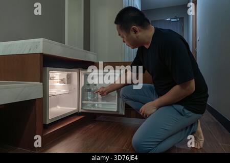 Ein Mann öffnet einen kleinen Kühlschrank, der in einem Holzschrank in einem dunklen Hotelzimmer eingebaut ist, und das warme Innenlicht steht im Kontrast zur dunklen Umgebung. Stockfoto