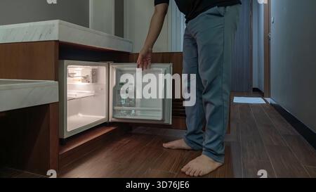 Ein Mann öffnet einen kleinen Kühlschrank, der in einem Holzschrank in einem dunklen Hotelzimmer eingebaut ist, und das warme Innenlicht steht im Kontrast zur dunklen Umgebung. Stockfoto