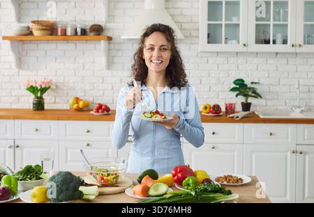 Lächelnde junge Frau genießt frischen Gemüsesalat in der Küche und fördert einen gesunden Lebensstil. Stockfoto