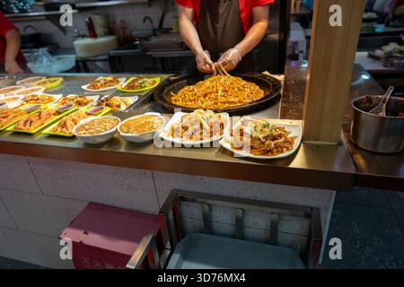 Ein Koch mit Handschuhen bereitet in einem großen schwarzen Wok in einem asiatischen Street Food-Stand gebratene Nudeln zu. Zubereitete Nudelgerichte und Suppenschüsseln sind Stockfoto