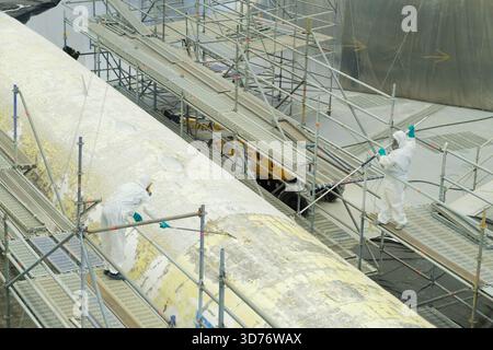 Zwei Arbeiter in Schutzanzügen reinigen einen großen Flugzeugflügel, der durch Gerüste in einem Hangar gestützt wird. Sie führen Wartungsaufgaben mit Werkzeugen und durch Stockfoto