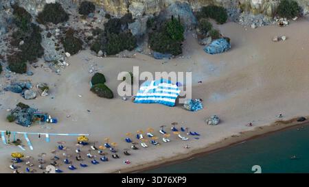 Aus der Vogelperspektive sehen Sie eine lebhafte griechische Flagge, die kühn auf dem goldenen Sand gemalt ist, im Kontrast zum azurblauen Meer und den sonnenverwöhnten Sonnenschirmen, Kolympia, Rod Stockfoto