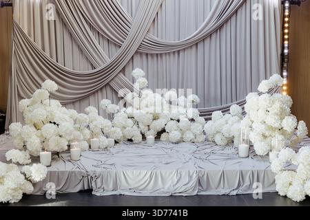 Eine atemberaubende Präsentation weißer Blumen und Kerzen ist wunderschön auf weichem, drapiertem Stoff angeordnet, perfekt für eine besondere Veranstaltung. Stockfoto
