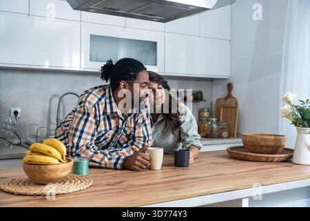 Fröhliches Paar, das in der Küche einen entspannten Kaffee-Chat genießt, sich auf die Theke lehnt, lächelt und über Pläne spricht, gemütliche Atmosphäre am Morgen Stockfoto