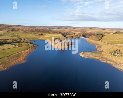 Aus der Vogelperspektive auf ein ruhiges Reservoir, das den Himmel spiegelt, umgeben von hügeligen Hügeln und Feldern in gedämpften Herbsttönen, befindet sich eine kleine bewaldete Insel Stockfoto