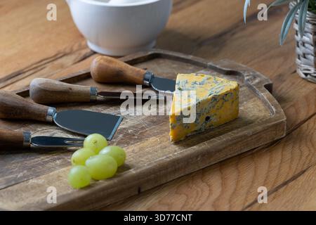 Blauschimmelkäse und verschiedene Käsemesser auf einer rustikalen Eichenkäseplatte mit Stößel und Mörtel und weichem floralem Hintergrund in natürlichem Fensterlicht angeordnet Stockfoto