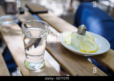 Kleines Glas Tequila mit Zitronenscheiben, serviert auf einem Holztisch in einem sonnigen Café im Freien Stockfoto