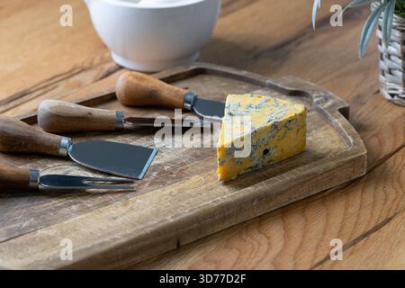 Blauschimmelkäse und verschiedene Käsemesser auf einer rustikalen Eichenkäseplatte mit Stößel und Mörtel und weichem floralem Hintergrund in natürlichem Fensterlicht angeordnet Stockfoto