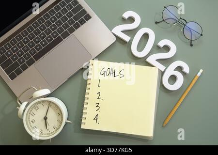 Flaches offenes Notebook mit der Aufschrift „Goals“, mit Auflistung der Dinge, die zu tun sind, Anzahl Neujahr 2026, Bleistift, Laptop, Wecker und Brille auf grünem Schreibtisch Stockfoto