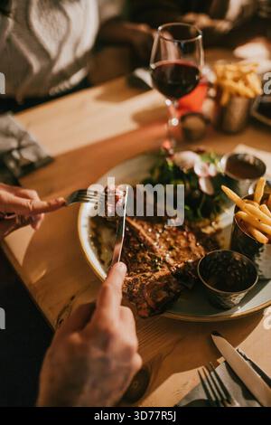 Eine Person isst ein Steak mit Messer und Gabel. Auf dem Teller gibt es Pommes und Salat. Die Atmosphäre des Bildes ist lässig und entspannt, wie die Stockfoto