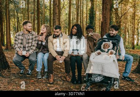 Eine Gruppe von Menschen sitzt in einem Wald, einer von ihnen schiebt einen Kinderwagen. Die Gruppe ist vielfältig, mit Menschen unterschiedlichen Alters und Rassen. Das sind sie Stockfoto