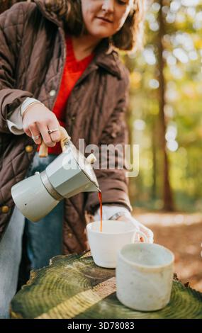Eine Frau gießt Kaffee aus einer Kaffeekanne in eine Tasse. Die Szene spielt in einem Wald mit Bäumen im Hintergrund. Die Frau trägt eine braune jacke Stockfoto