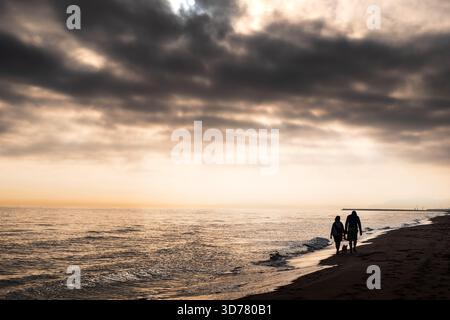 Ein paar Spaziergänge am Gandia Sandstrand in der Abenddämmerung, begleitet von einem verspielten kleinen Hund. Sanftes Licht der untergehenden Sonne reflektiert auf sanften Wellen Stockfoto