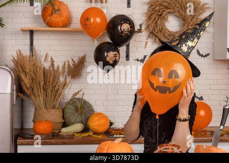 Eine unerkennbare Frau, die ein gruseliges Halloween-Gesicht vor ihrem Gesicht hält, einen leuchtend orangen Ballon. Eine Frau in einem Karnevalshut versteckt ihr Gesicht hinter einer Kürbislaterne. Hintergrund für Halloween-Weihnachtsdekoration Stockfoto