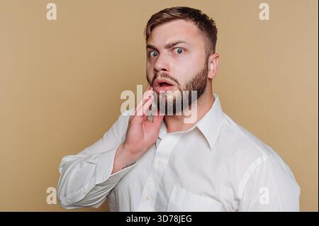 Porträt eines jungen Mannes mit überraschtem Ausdruck, der sein Gesicht auf beigefarbenem Hintergrund hält Stockfoto