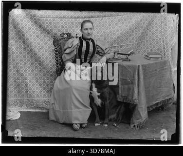 Amsterdam, 1895: Das historische Schwarz-weiß-Atelierporträt von Jacob Olie zeigt eine Frau in viktorianischem Kleid, die neben einem Tisch mit Büchern sitzt und ihre Hand auf einem großen Hund an ihrer Seite hält. Die Szene zeigt eine gemusterte Kulisse, einen kunstvollen Stuhl und eine drapierte Tischdecke, die die Atmosphäre und die häusliche Ästhetik der niederländischen Porträtfotografie des späten 19. Jahrhunderts einfangen. Ein detaillierter Einblick in den Alltag und die Studiokultur Amsterdams der 1890er Jahre Stockfoto