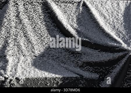 Eine graue Kunststoffplane, die mit einer Schneeschicht bedeckt ist, erzeugt ein kaltes Wintermuster mit einer rauen Textur und weichen Lichtkontrasten Stockfoto